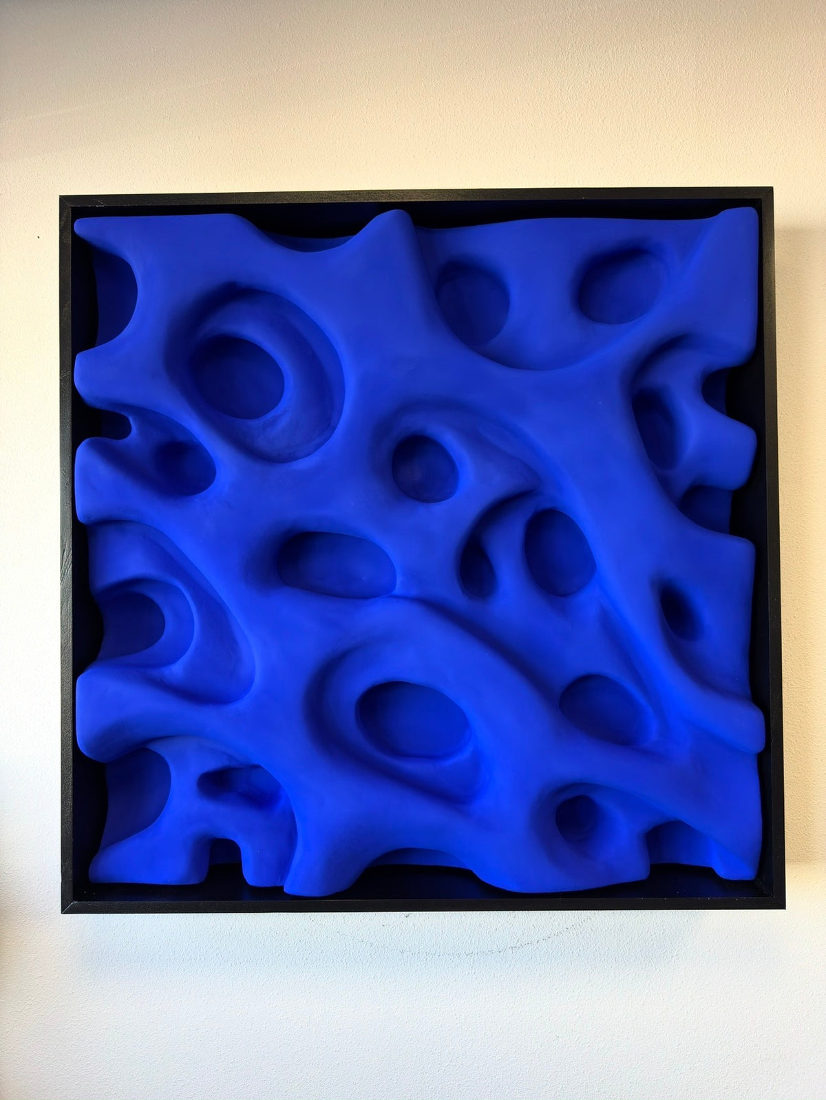 Alt text: Square matte Yves Klein Blue abstract relief artwork with organic recessed shapes in a black shadowbox frame, 105 × 105 cm.
